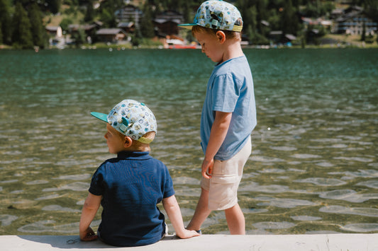 Two young boys standing by a lake, wearing matching patterned snapback caps, light blue and navy shirts, and light-colored shorts. The background shows a scenic outdoor lake with trees and houses on the shore.