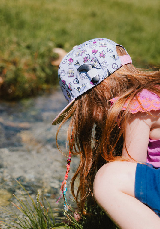 A child wearing a light purple snapback cap covered in colorful illustrations and geometric patterns, photographed outdoors near a stream.