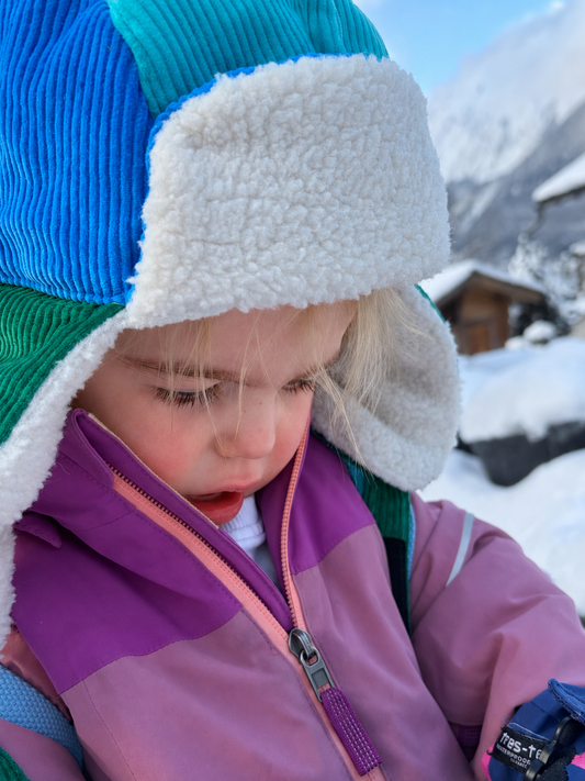 A close-up lifestyle shot of a young child wearing a colorful winter outfit. The outfit features an ear flap hat in blue, green, and teal with a white fleece lining, and a color-blocked purple and pink ski jacket.