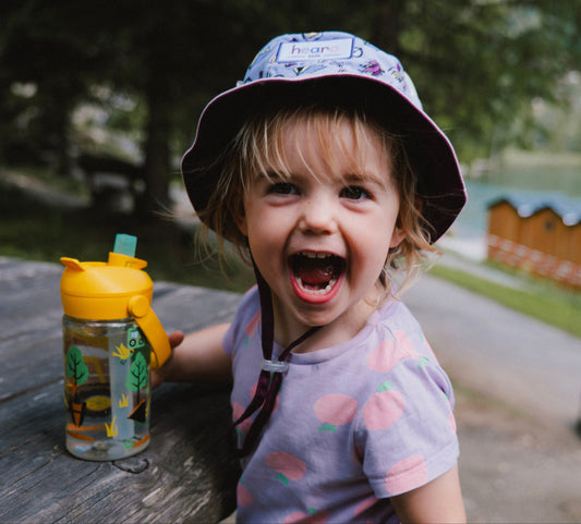 A young child wearing a patterned bucket hat and a light purple t-shirt with pink fruit prints, smiling broadly while leaning on a wooden picnic table next to a yellow and clear water bottle. The background shows a blurred outdoor lakeside setting with wooden cabins.
