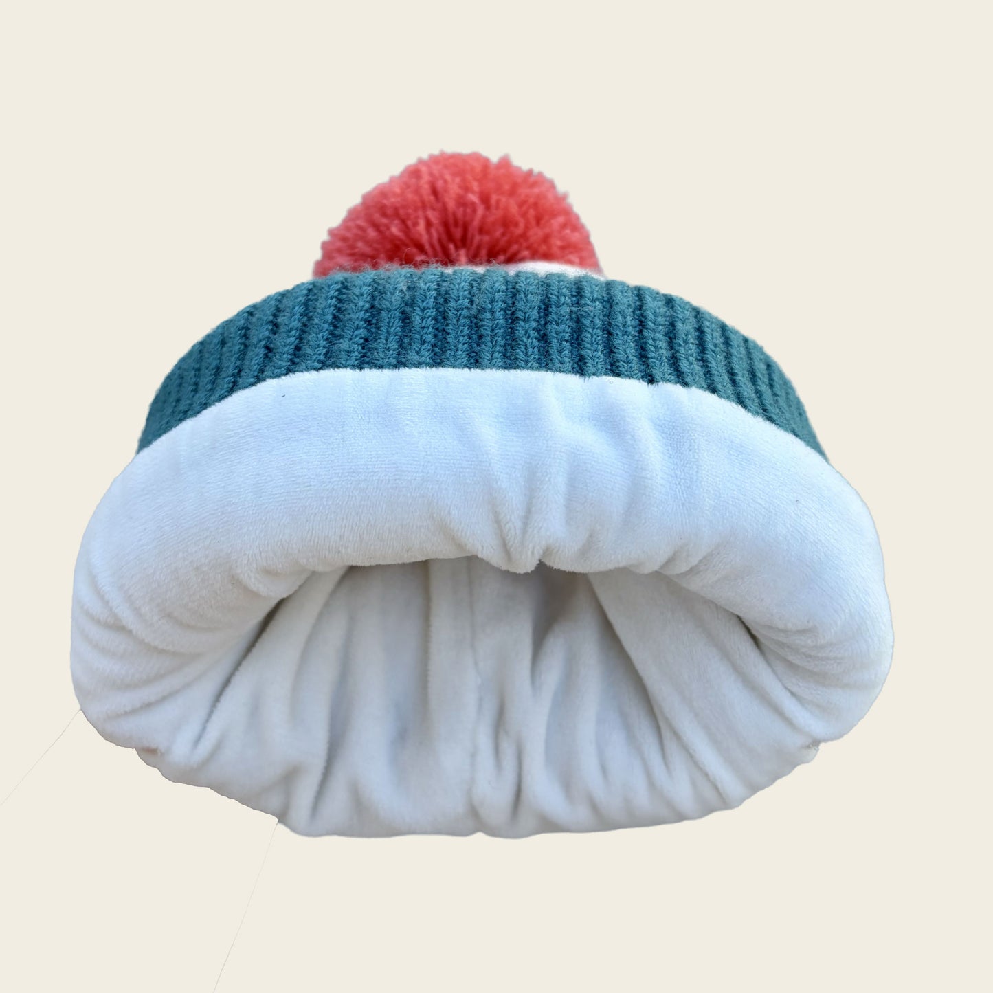 A folded knit beanie hat with a red pom-pom on top, featuring a dark teal ribbed band and a thick white fleece or plush inner layer, shown against a plain light beige background.