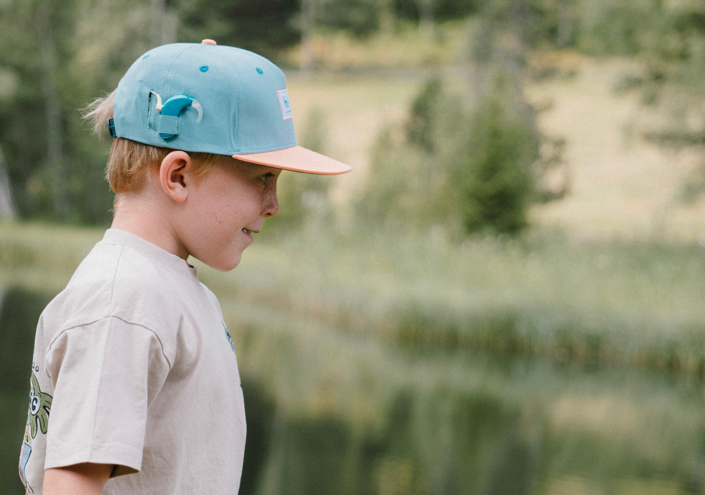 A young boy in profile wearing a light teal green snapback cap with a peach brim and a beige t-shirt, standing outdoors in front of a blurred lake and forest background.