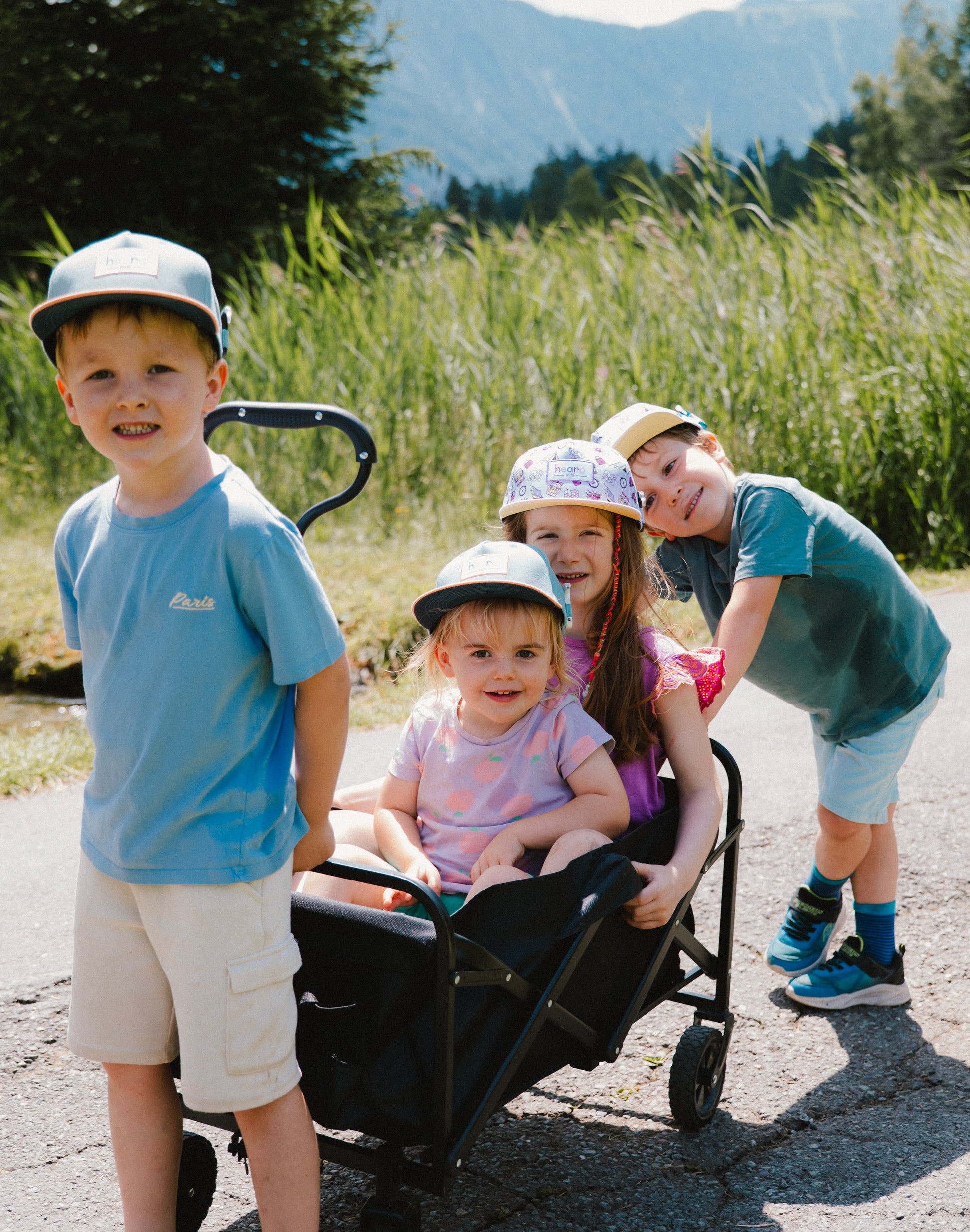 Four children outdoors on a sunny day, with three wearing matching light blue and green baseball caps while pushing and sitting in a black wagon on a paved path with tall grass and mountains in the background.