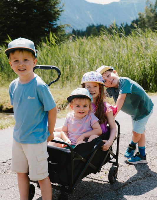 Four children outdoors on a sunny day, with three wearing matching light blue and green baseball caps while pushing and sitting in a black wagon on a paved path with tall grass and mountains in the background.