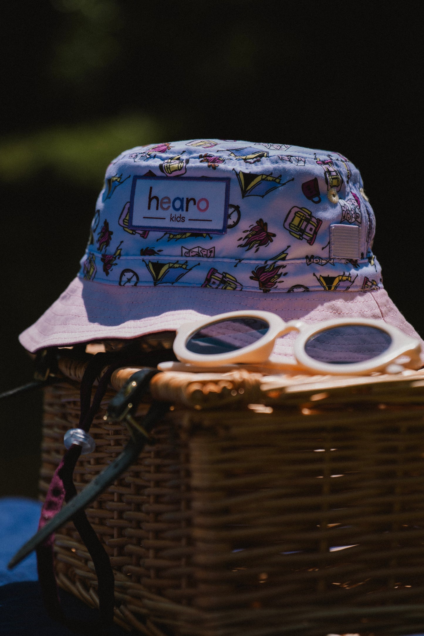 A light purple patterned bucket hat with a visible 'hearo' label, resting on top of a wicker picnic basket with white-framed sunglasses on a dark outdoor background.