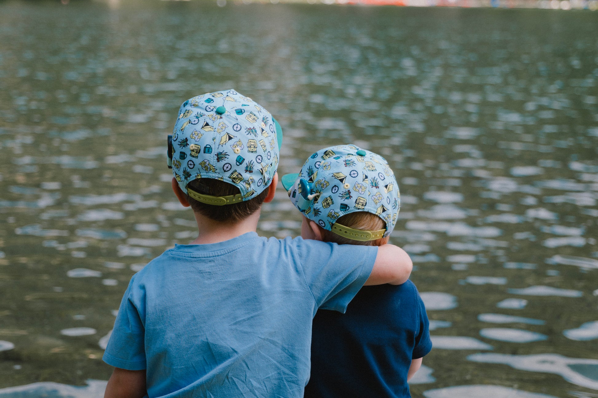 Two children seen from behind wearing matching light blue patterned snapback caps and blue shirts, standing by a body of water.