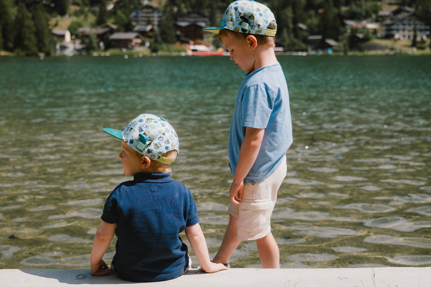 Two young boys standing by a lake, wearing matching patterned snapback caps, light blue and navy shirts, and light-colored shorts. The background shows a scenic outdoor lake with trees and houses on the shore.