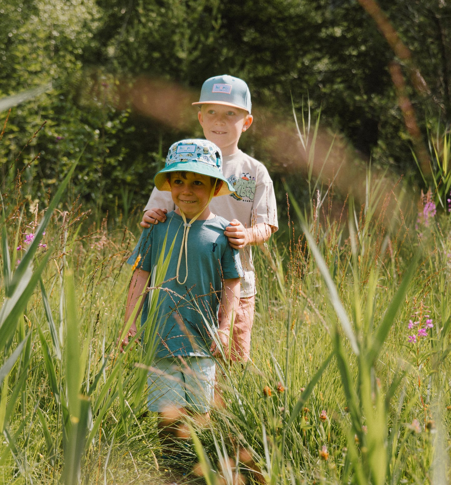 Two children walking through tall grass outdoors, one wearing a bucket hat and the other a snapback cap. The child in front wears a blue t-shirt, and the child behind wears a beige t-shirt.