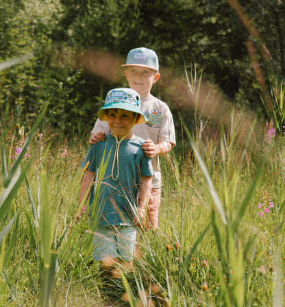 Two children walking through tall grass outdoors, one wearing a bucket hat and the other a snapback cap. The child in front wears a blue t-shirt, and the child behind wears a beige t-shirt.