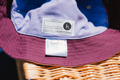 Close-up of a children's sun hat with a maroon brim and light purple inner lining, resting on a wicker basket. The brand label reads 'hearo kids'.