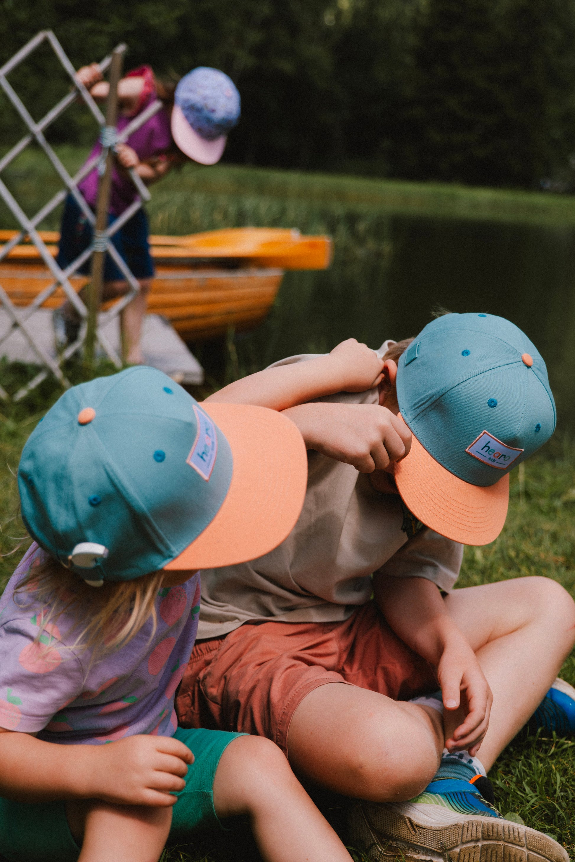 Two children sitting on grass by a lake wearing matching teal and peach snapback caps, with a third child and a wooden boat visible in the background.