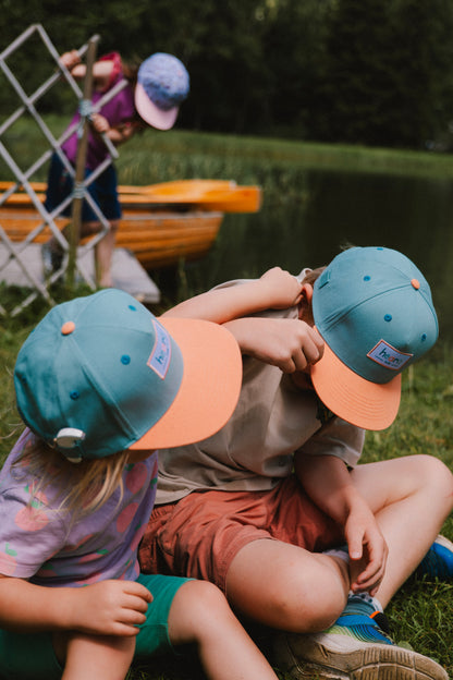 Two children sitting on grass by a lake wearing matching teal and peach snapback caps, with a third child and a wooden boat visible in the background.