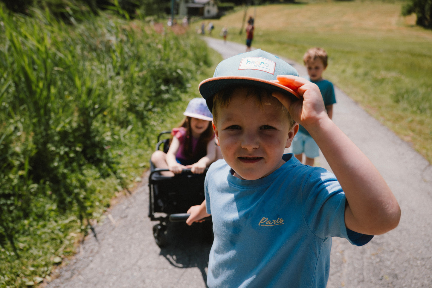 A young boy in a blue t-shirt and a teal snapback cap with an orange brim smiles while pushing a stroller on a paved path outdoors, with other children behind him in a sunny park setting.