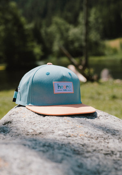 A light teal snapback cap with a peach-colored flat brim and a rectangular 'hearo' patch on the front, resting on a large gray rock with a blurred outdoor forest background.