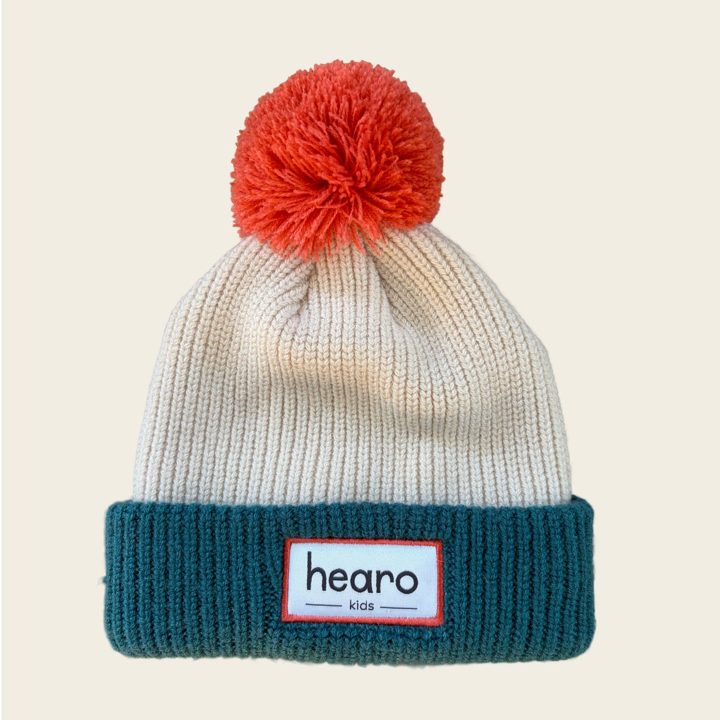 A ribbed knit beanie hat with a large orange-red pom-pom on top, a cream upper section, and a dark teal folded cuff. A white rectangular patch on the cuff reads 'hearo kids'.
