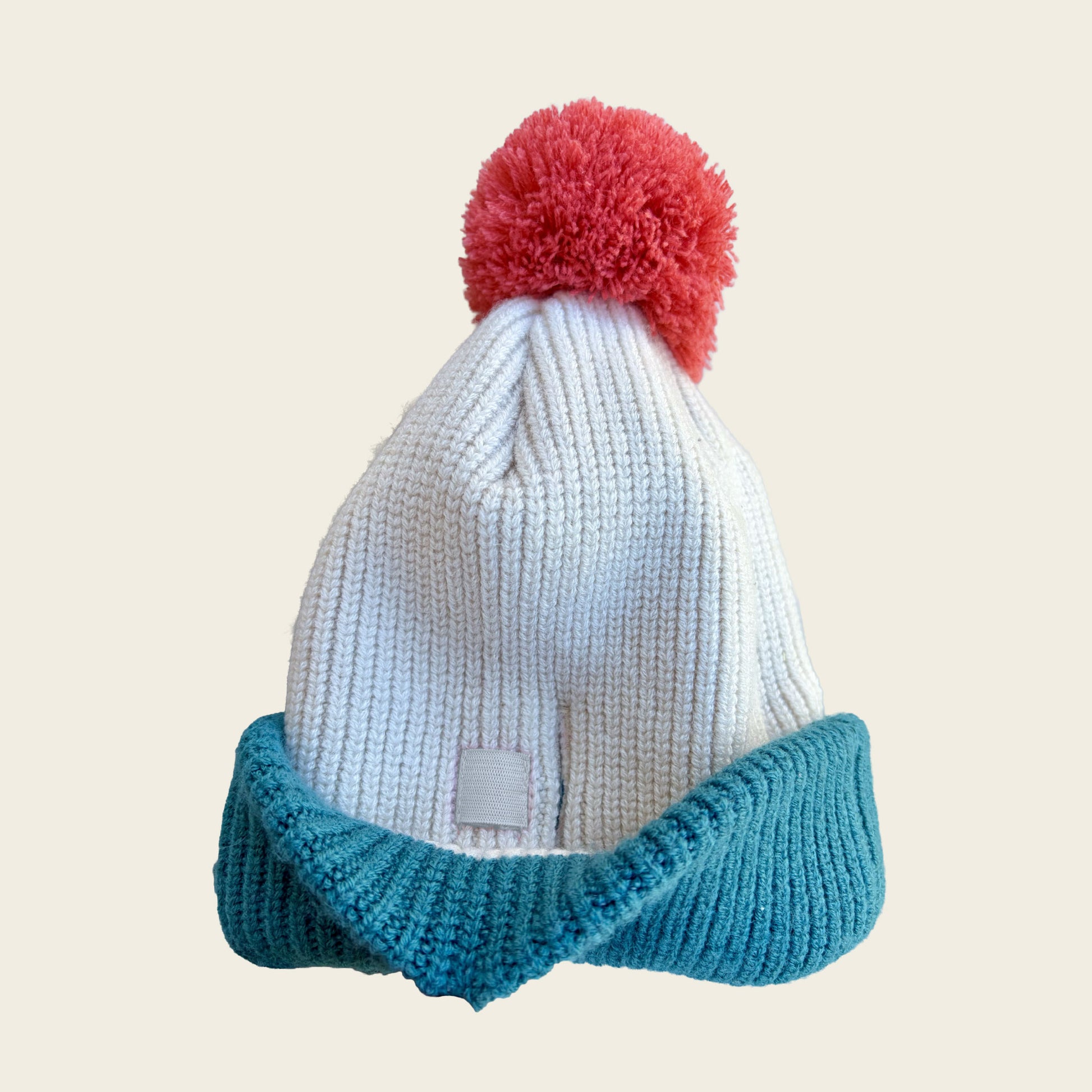A ribbed knit beanie hat featuring a white crown, a teal blue folded cuff, and a red pom-pom on top, shown against a solid light beige background.