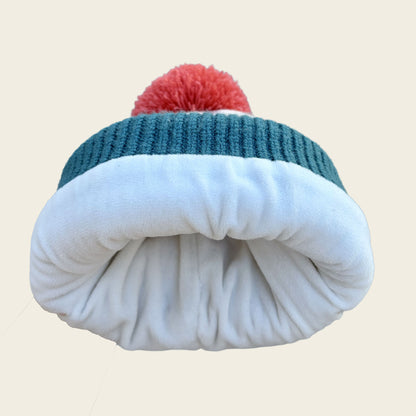 A folded knit beanie hat with a red pom-pom on top, featuring a dark teal ribbed band and a thick white fleece or plush inner layer, shown against a plain light beige background.