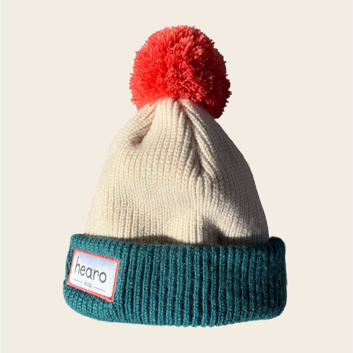 A knit beanie hat with a cream-colored upper section, a teal ribbed cuff, and a bright red pom-pom on top. A white sewn-on label on the cuff reads 'hearo'.