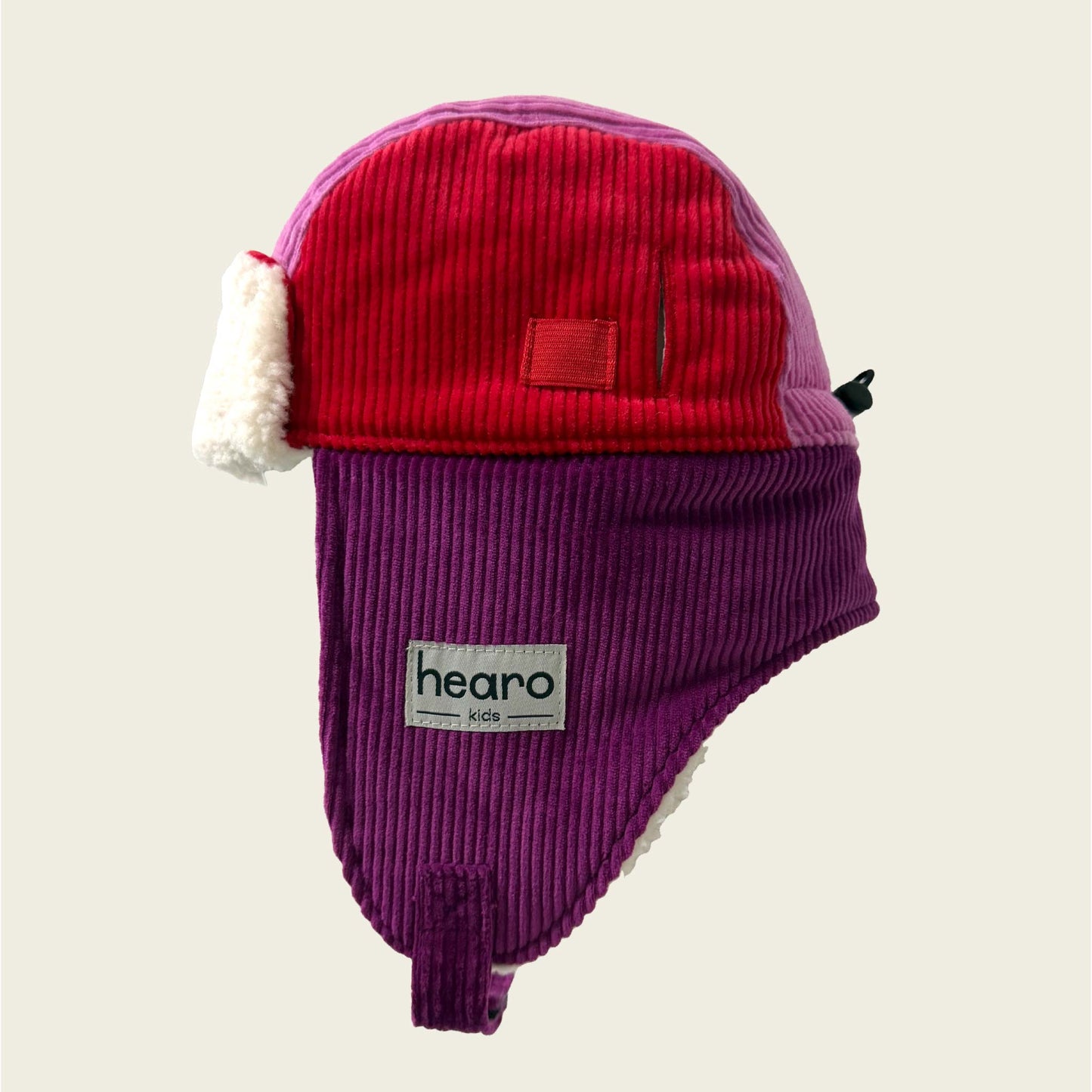 Two-tone red and purple children's ear flap hat with a visible brand logo on a beige background