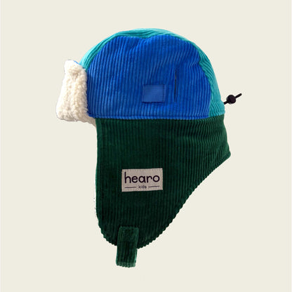 Two-tone blue and green hat with white earflaps on a beige background, featuring the 'hearo' brand.