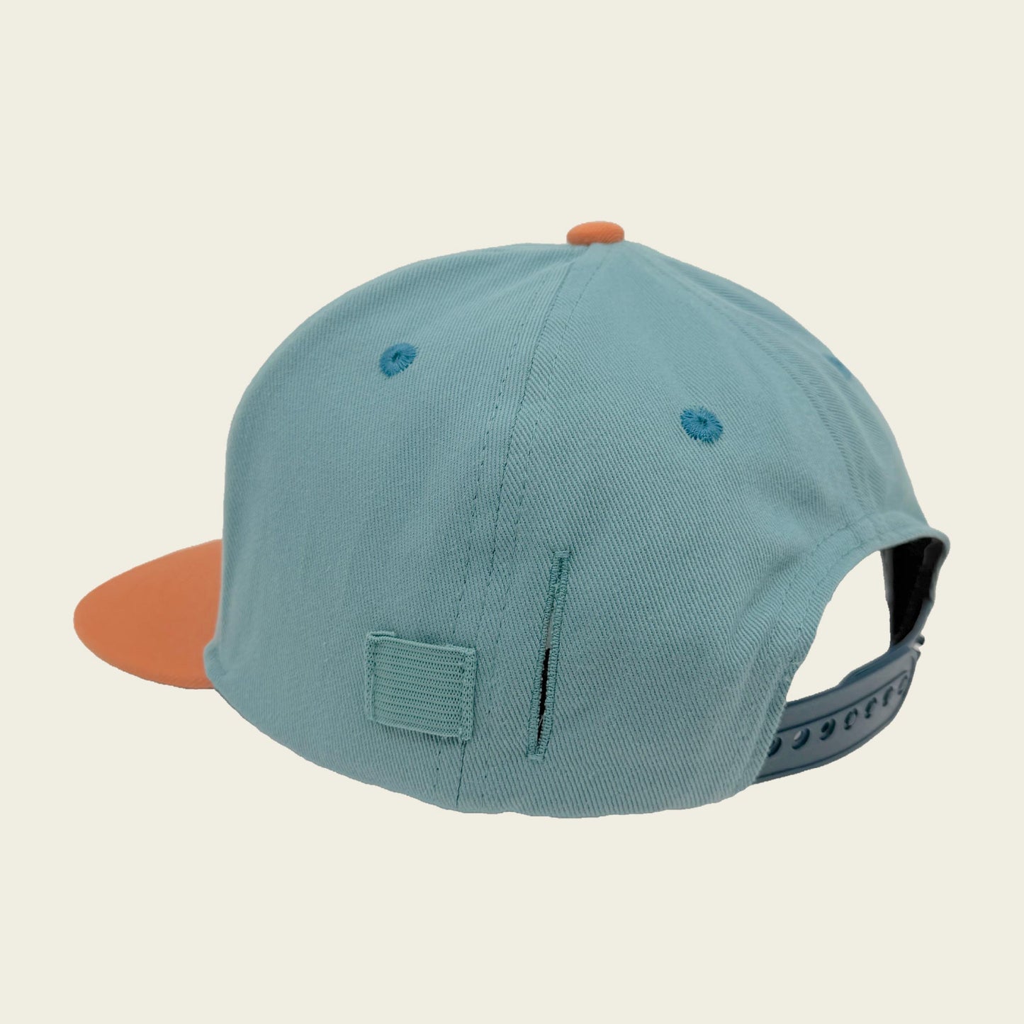 A light teal green snapback cap with a contrasting orange visor and orange button, shown from a three-quarters back angle on a light beige background.