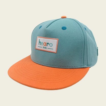 A two-tone snapback cap for kids with a light teal crown and a bright orange flat brim. The front features a white rectangular patch with the text 'hearo kids' in orange and teal.
