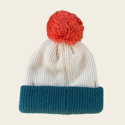 A ribbed knit beanie hat with a cream-colored upper section, a teal cuffed bottom, and a bright red-orange pom-pom on top, shown against a solid light beige background.