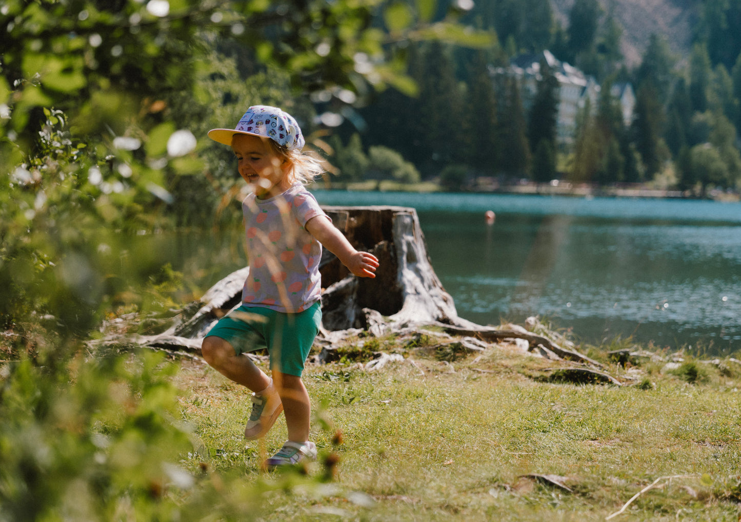A young child wearing a purple patterned cap, a light pink top, and teal shorts runs along a grassy lakeshore with trees and water in the background.