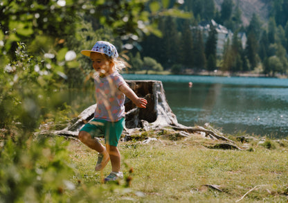 A young child wearing a purple patterned cap, a light pink top, and teal shorts runs along a grassy lakeshore with trees and water in the background.