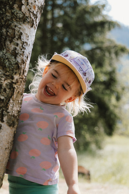 A young child wearing a light purple short-sleeve t-shirt with peach prints and a patterned cap, leaning against a tree outdoors.